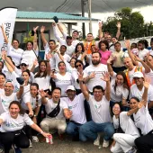 A large group of volunteers from AES Panamá pose together outdoors, smiling and raising their hands in celebration. They wear white Fundación AES Panamá shirts, with some in safety vests, standing in front of a building and AES banners. The group exudes enthusiasm and unity, showcasing their community involvement and volunteer spirit.