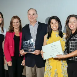 Five individuals pose together at an award ceremony. A man in a suit and four women dressed elegantly stand in front of a plain backdrop. Two women hold a certificate and a trophy, representing recognition received by Fundación AES Panamá. All individuals smile, showcasing a celebratory and formal atmosphere.