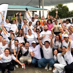 A large group of volunteers from AES Panamá pose together outdoors, smiling and raising their hands in celebration. They wear white Fundación AES Panamá shirts, with some in safety vests, standing in front of a building and AES banners. The group exudes enthusiasm and unity, showcasing their community involvement and volunteer spirit.